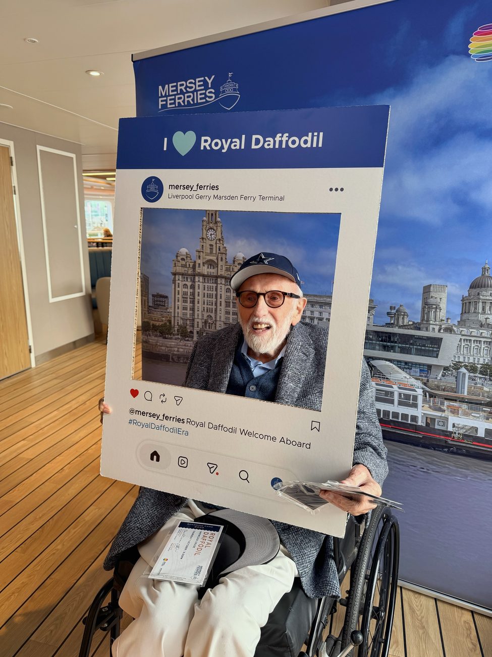 Mike Lowe in his wheelchair aboard the new Royal Daffodil, smiling through a giant cut-out frame that reads I love Royal Daffodil, with his boarding card for the showcase cruise on his knee.