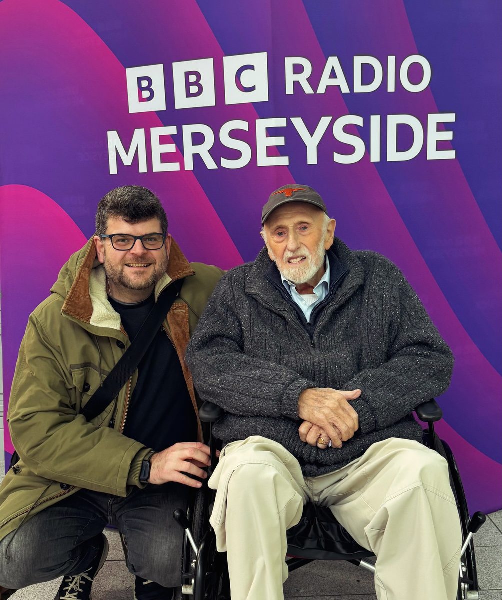 Rory Wilmer crouching beside Mike Lowe in his wheelchair in front of a BBC Radio Merseyside banner.